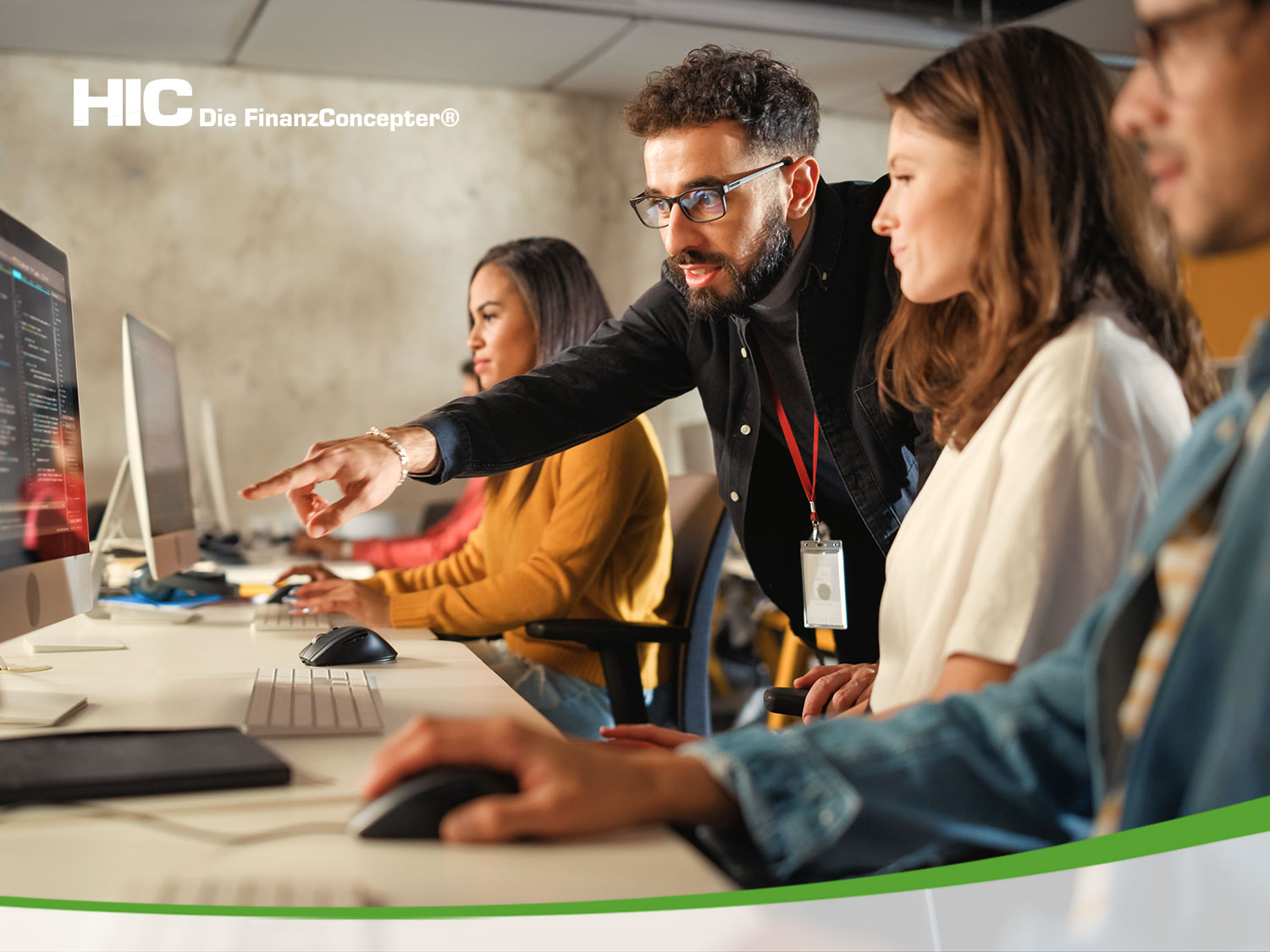 Mehreren Menschen sitzen an Computern. Ein Mann steht dahinter und deutet erklärend auf einen der Monitore.